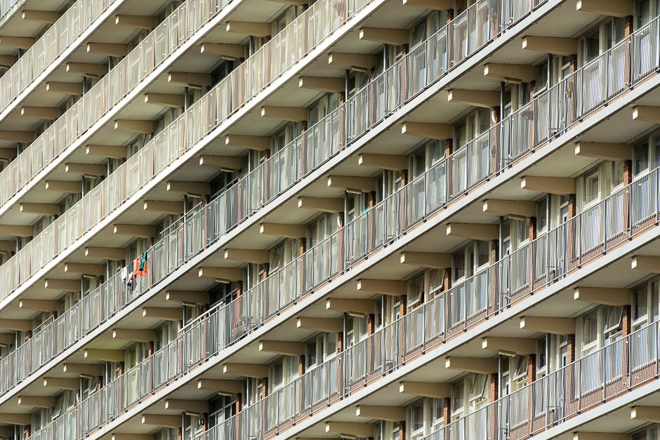 Front exterior view of a multi-story residential building with pink panel cladding, beige brick accents, and white-framed windows. Several apartment balconies with metal railings are visible, some fea