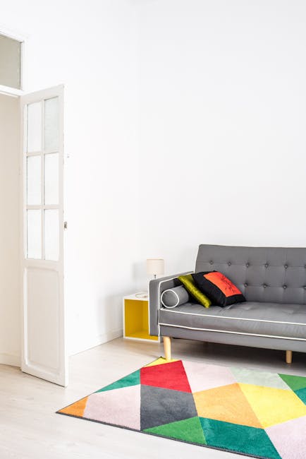 A modern living room with white walls and light wooden flooring featuring a colorful geometric patterned rug in shades of red, orange, yellow, green, and gray. A gray upholstered sofa with tufted butt